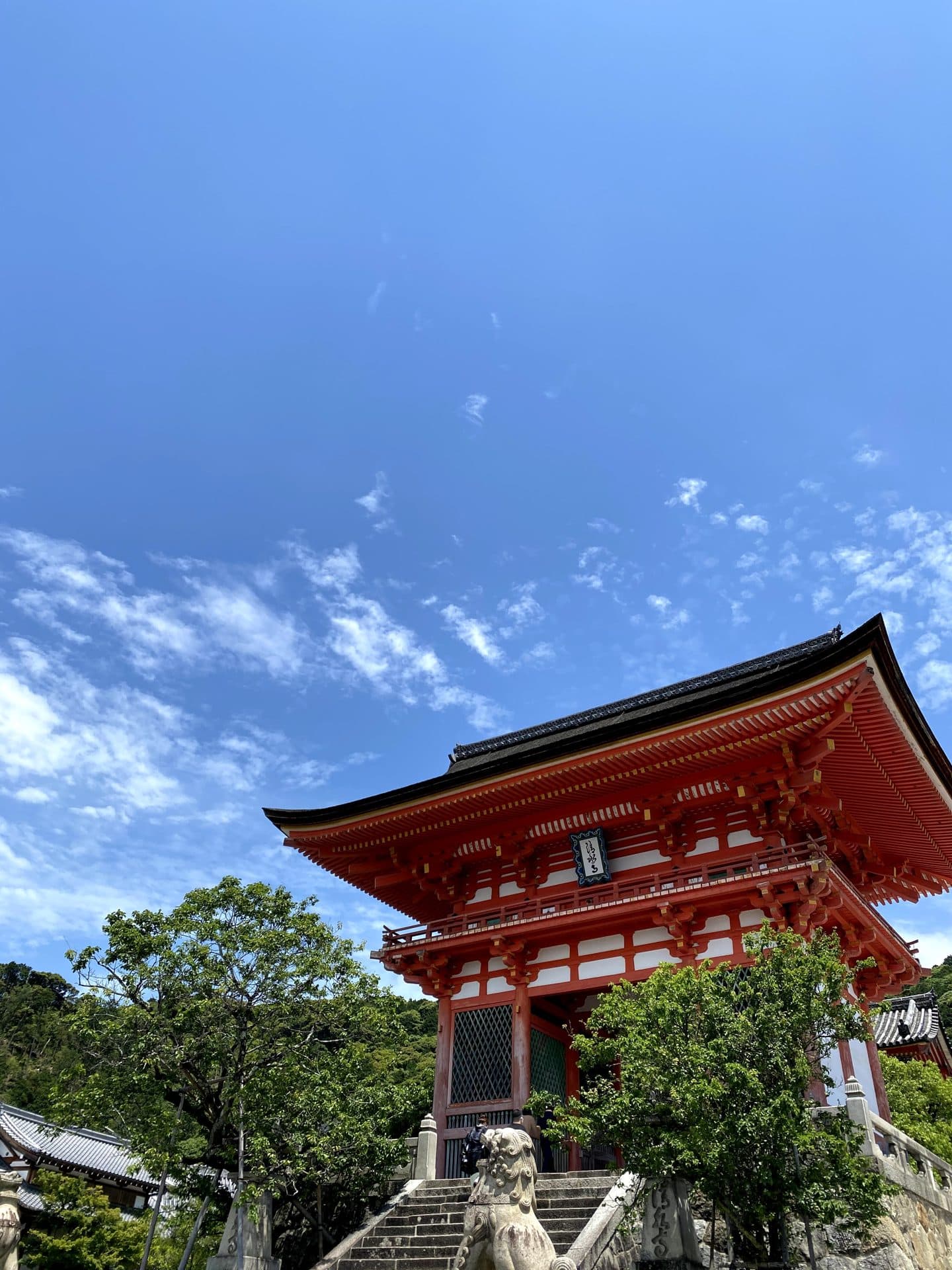 The vermilion Niomon gate of Kiyomizu-dera under a blue sky, with green hillside behind it