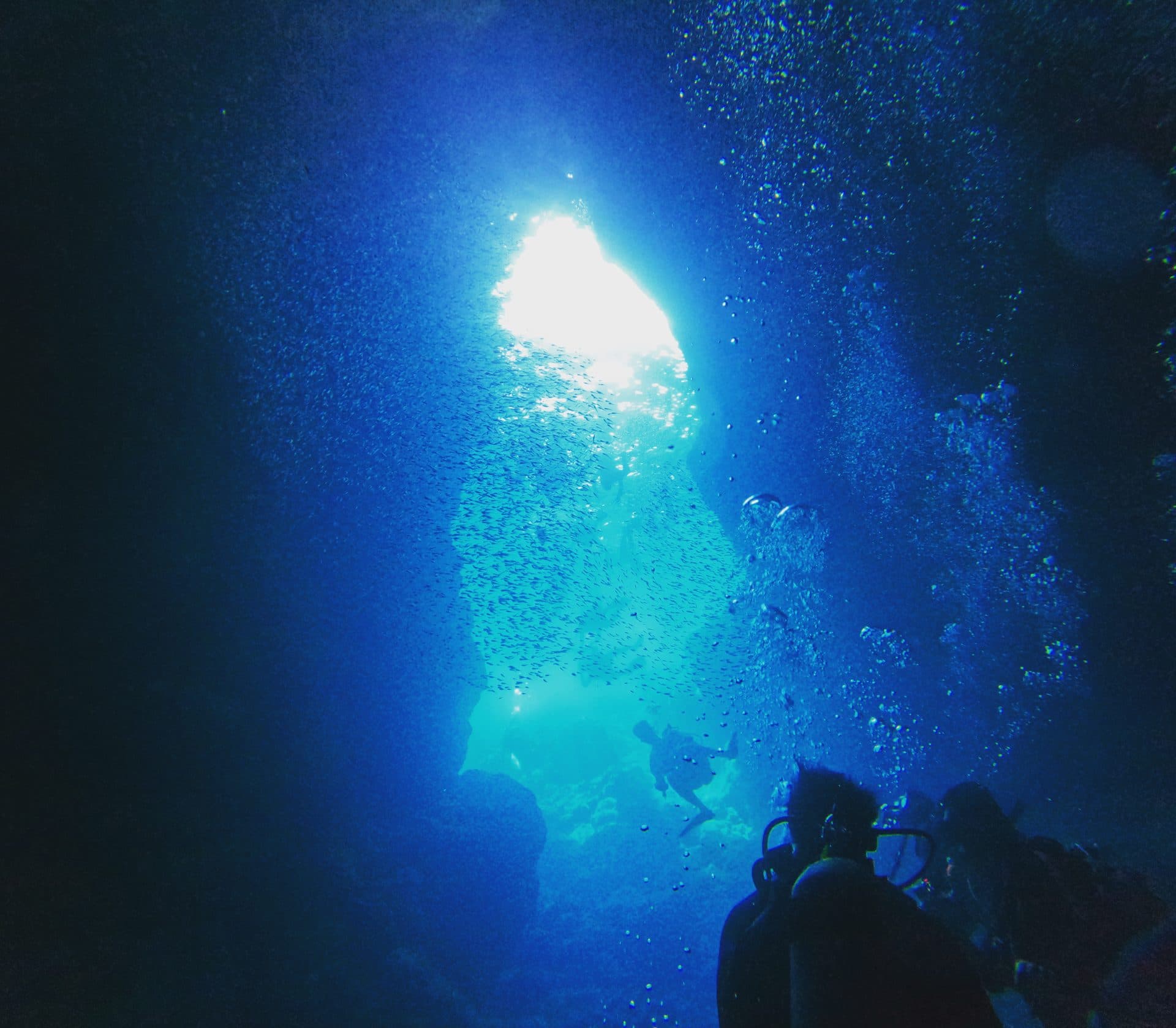 Divers silhouetted inside Okinawa's Blue Cave as sunlight streams through the cave mouth and illuminates a school of fish in electric blue water