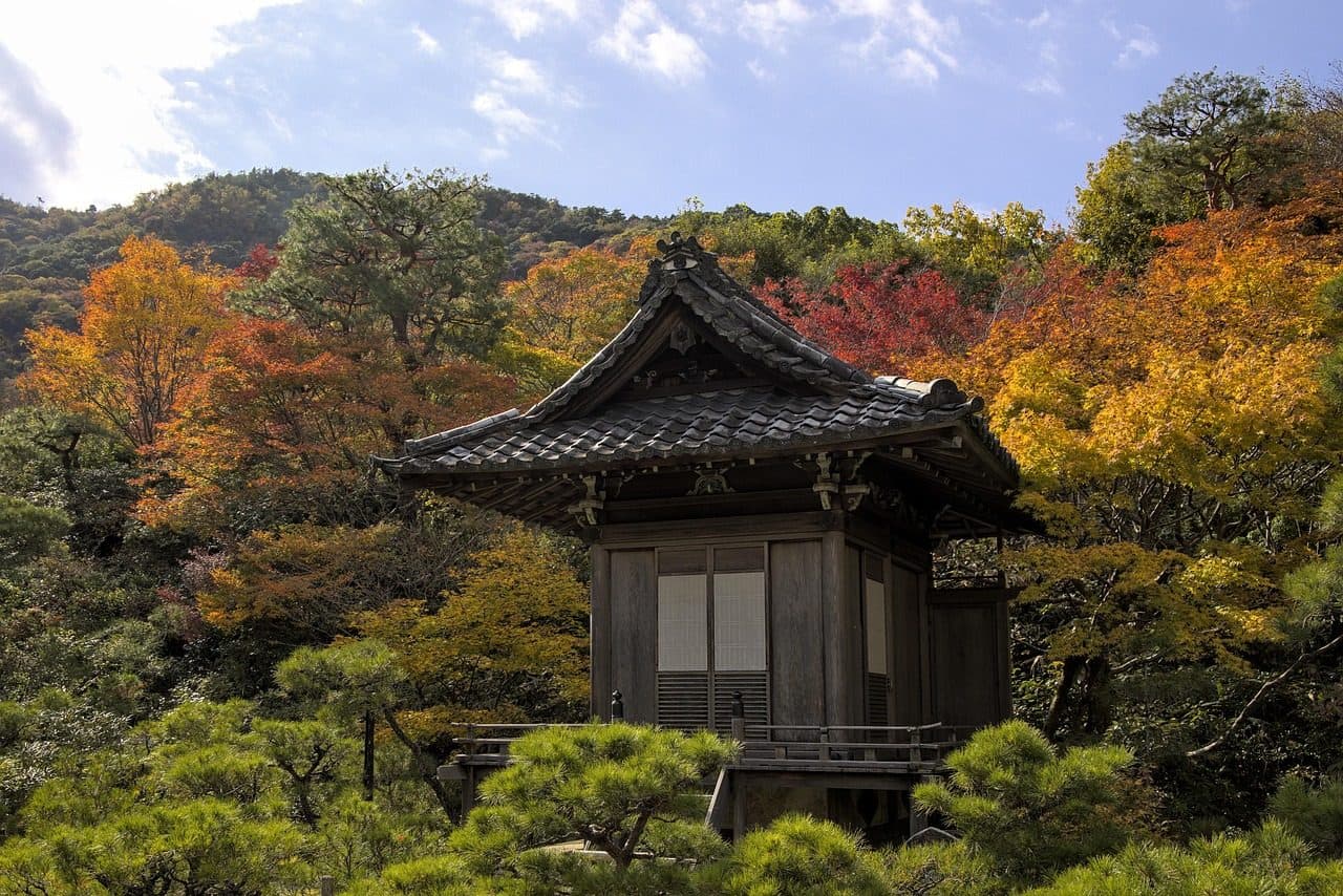 A small wooden temple hall in a Kyoto garden surrounded by red, orange and yellow autumn trees under a blue sky