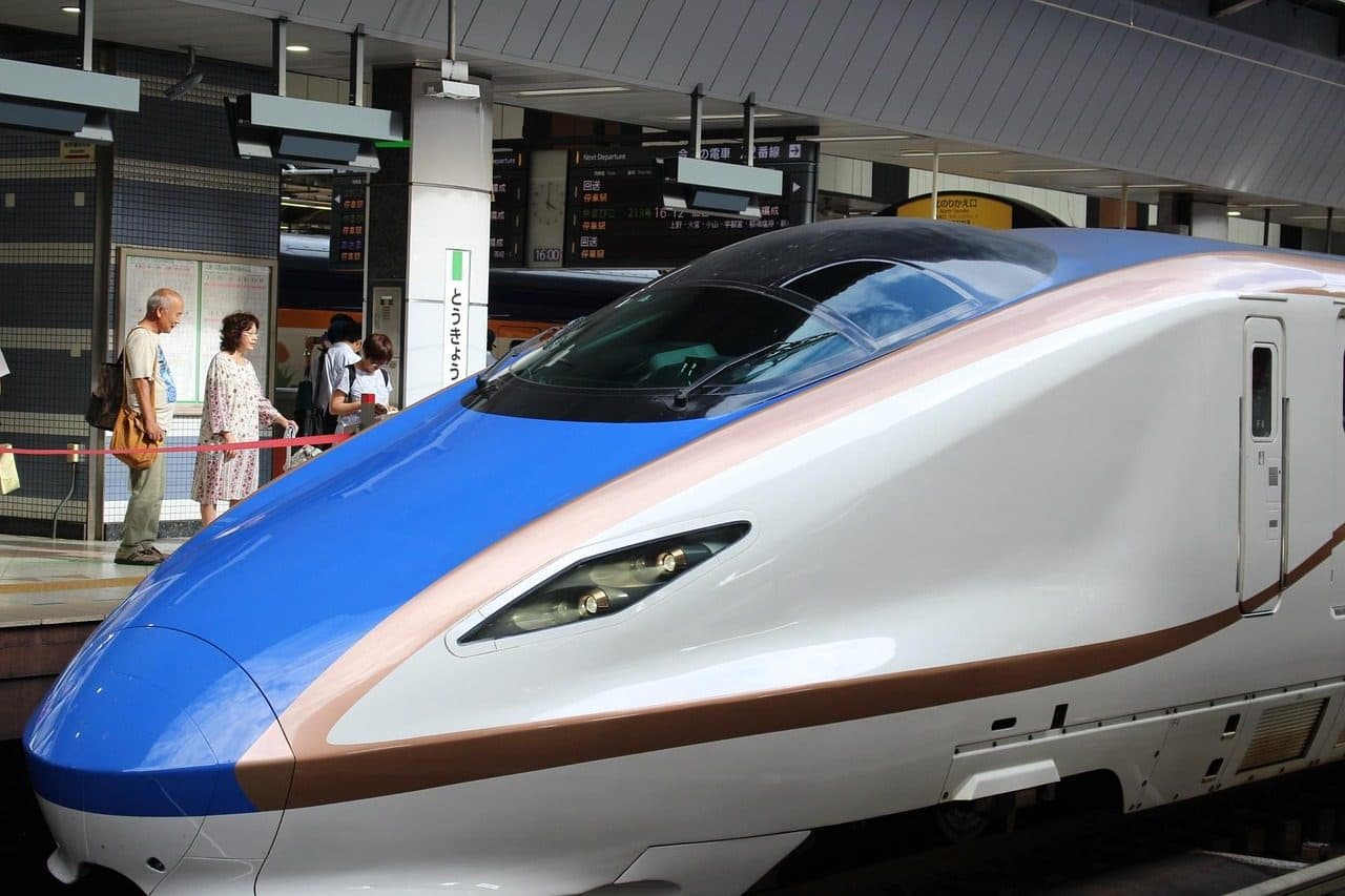 The blue, copper and white nose of an E7-series Hokuriku Shinkansen train at a Tokyo Station platform, with passengers waiting behind