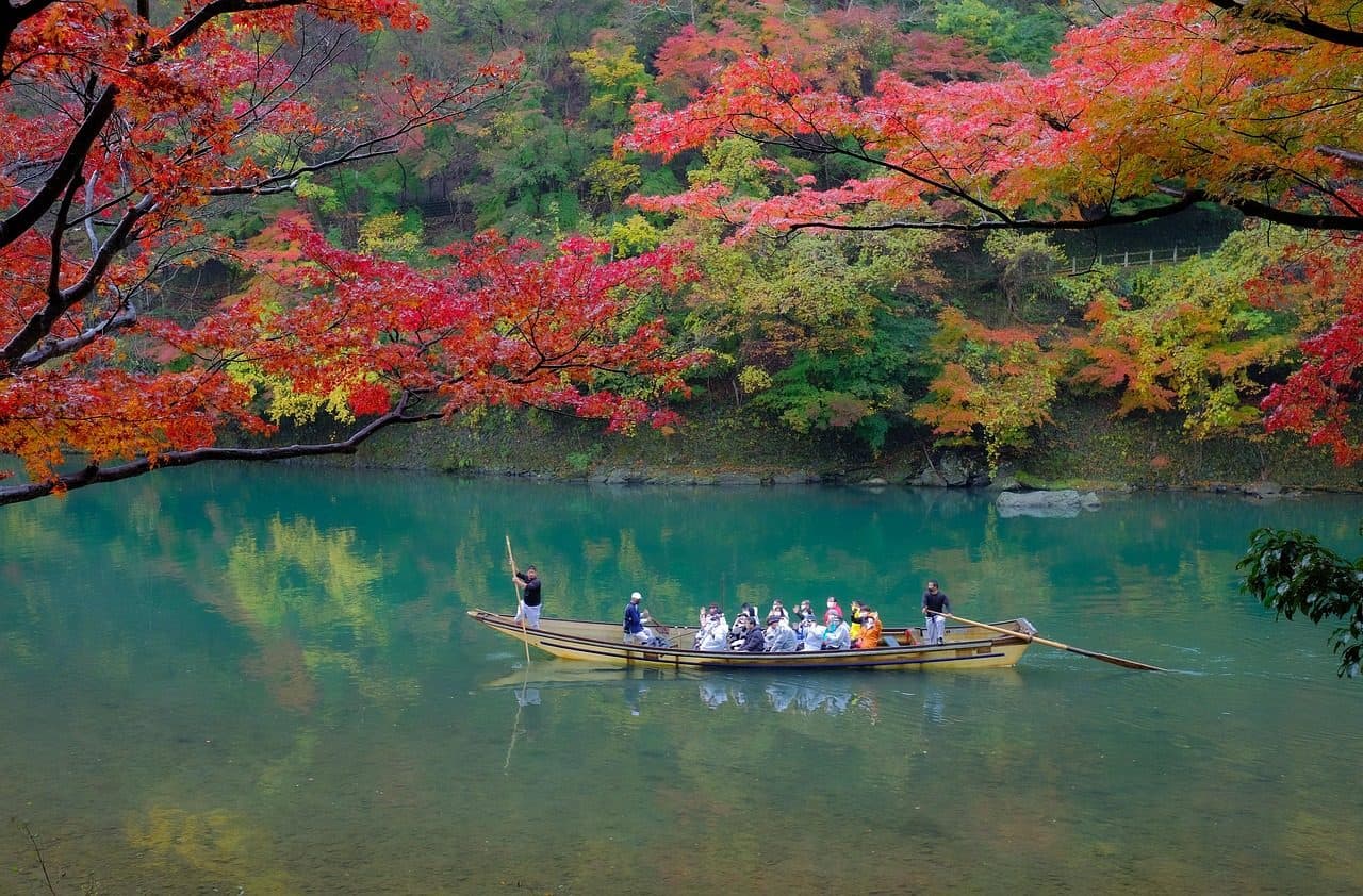 A wooden boat on the emerald Hozu River in Arashiyama, Kyoto, beneath red and orange autumn maples