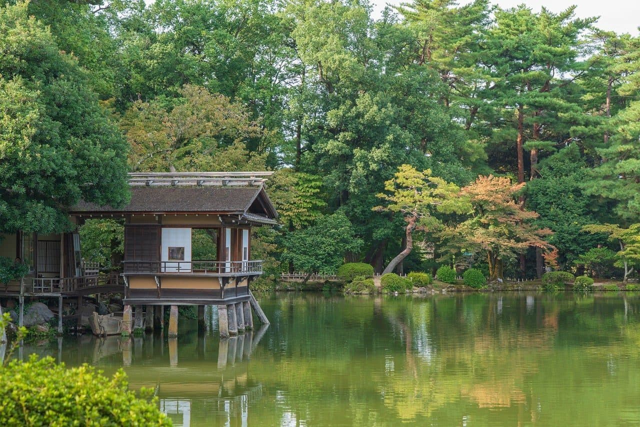 A wooden teahouse on stilts over a green pond surrounded by pine trees and early autumn foliage in Kenrokuen garden, Kanazawa