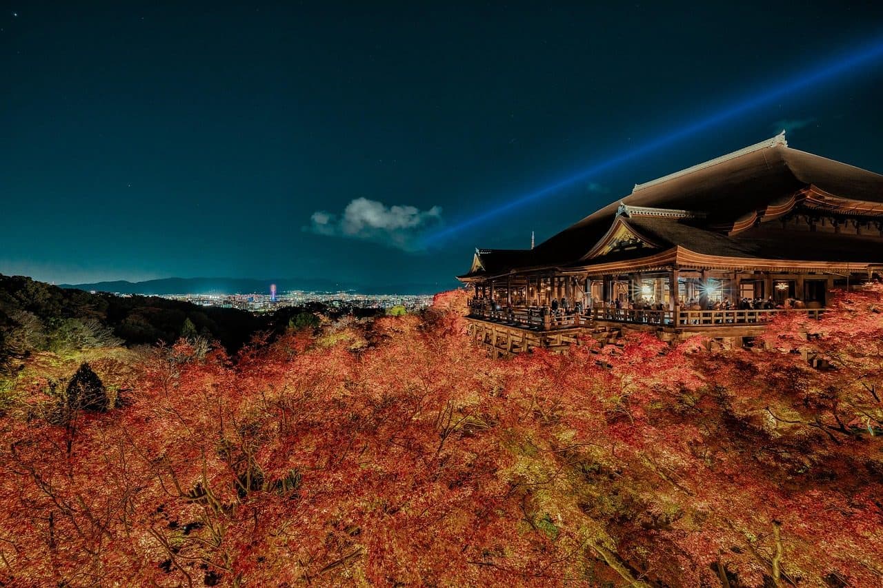 Kiyomizu-dera's main hall lit at night above a hillside of red maples, with a blue searchlight crossing the sky over Kyoto