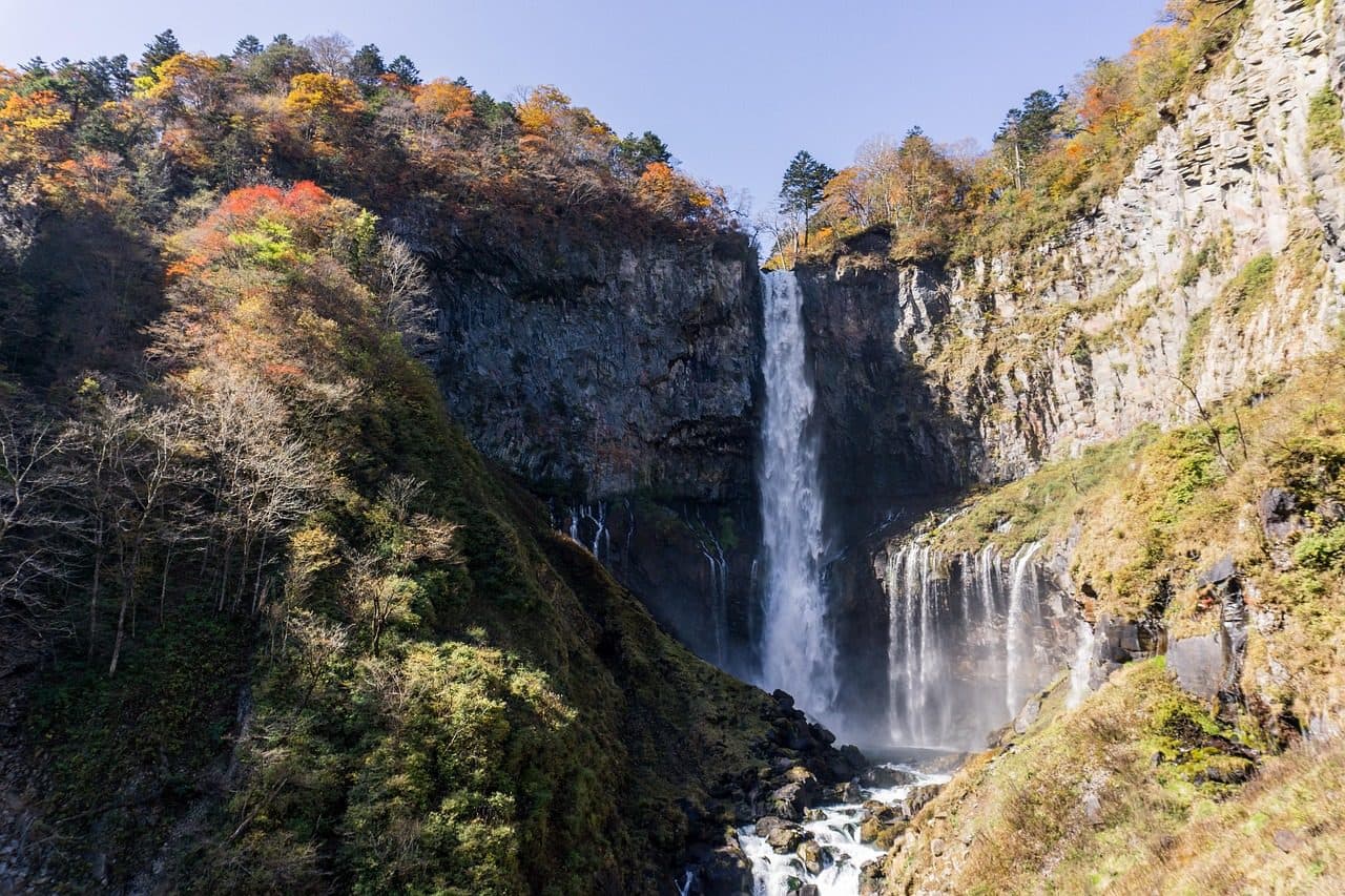 Kegon Falls in Nikko dropping down a dark rock cliff, with autumn-colored trees on the slopes above and smaller falls to the right