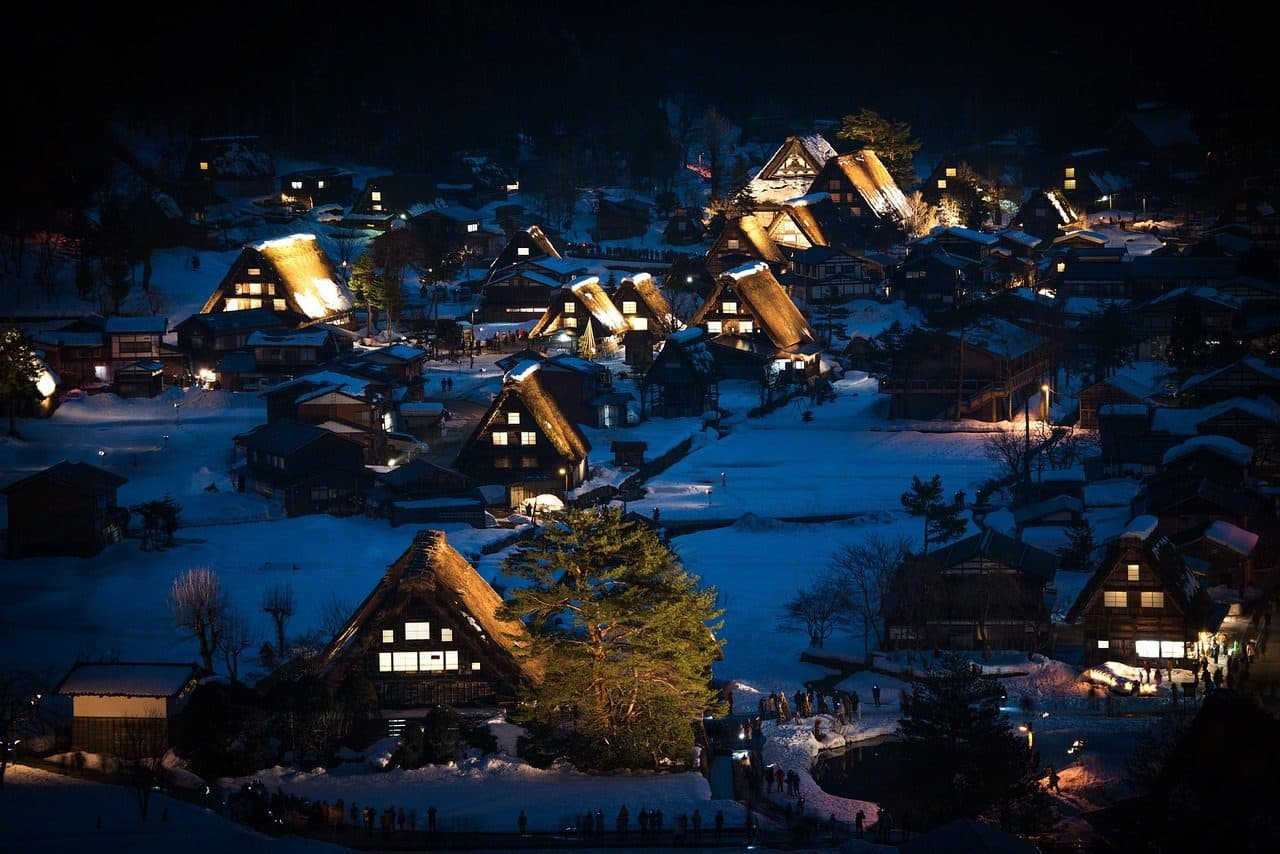 Shirakawa-go's thatched gassho-zukuri farmhouses lit up at night in deep winter snow, seen from above, with visitors walking the lanes